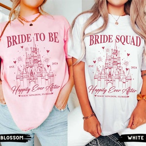 K&ouml;nnte beinhalten: Zwei wei&szlig;e T-Shirts mit einem rosa und wei&szlig;en Design von Cinderellas Schloss. Der Text auf den Shirts lautet "Bride to Be" und "Bride Squad" mit den Worten "Happily Ever After" und "Magic Kingdom, Florida" unterhalb des Schlosses. Die Shirts werden von Models getragen, die Jeans tragen.