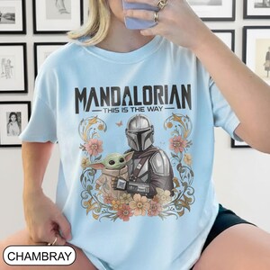 May include: Light blue t-shirt featuring the text "MANDALORIAN - THIS IS THE WAY" above an illustration of the Mandalorian and Grogu surrounded by flowers. The shirt is made of chambray fabric.