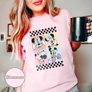May include: Pink t-shirt featuring a checkered pattern and four images of Minnie Mouse in various poses. The images are arranged in a grid pattern, each with a different coloured background.