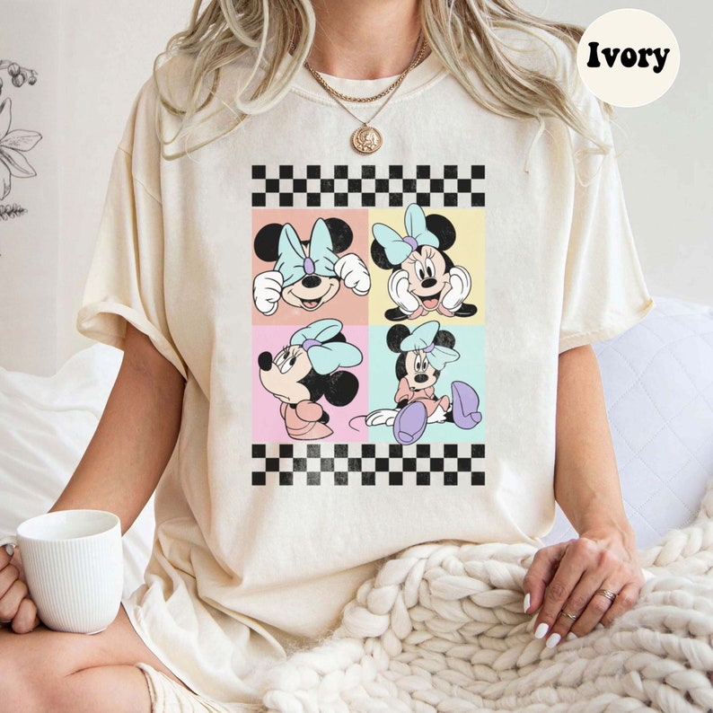 May include: A white t-shirt featuring a checkered pattern and a collage of four images of Minnie Mouse in various poses. The images are in a pastel colour palette and have a vintage look. The t-shirt is labelled "Ivory".