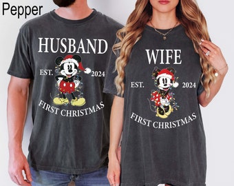 May include: Two matching dark gray t-shirts. One says "HUSBAND" and the other says "WIFE" in white text. Both shirts feature a Mickey Mouse graphic in a Santa hat, with the text "EST. 2024 FIRST CHRISTMAS".