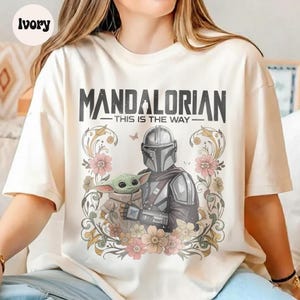 May include: Ivory-colored t-shirt featuring a graphic of The Mandalorian and Grogu surrounded by floral elements. The text "MANDALORIAN - THIS IS THE WAY" is printed above the characters. The design includes a butterfly and various pink and beige flowers.