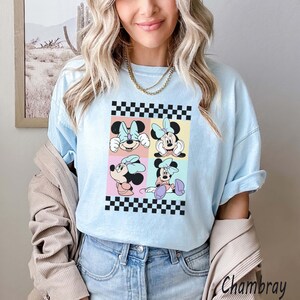 May include: A light blue t-shirt with a checkered border and a graphic of four different images of Minnie Mouse. The images are in a grid pattern and each image has a different coloured background.