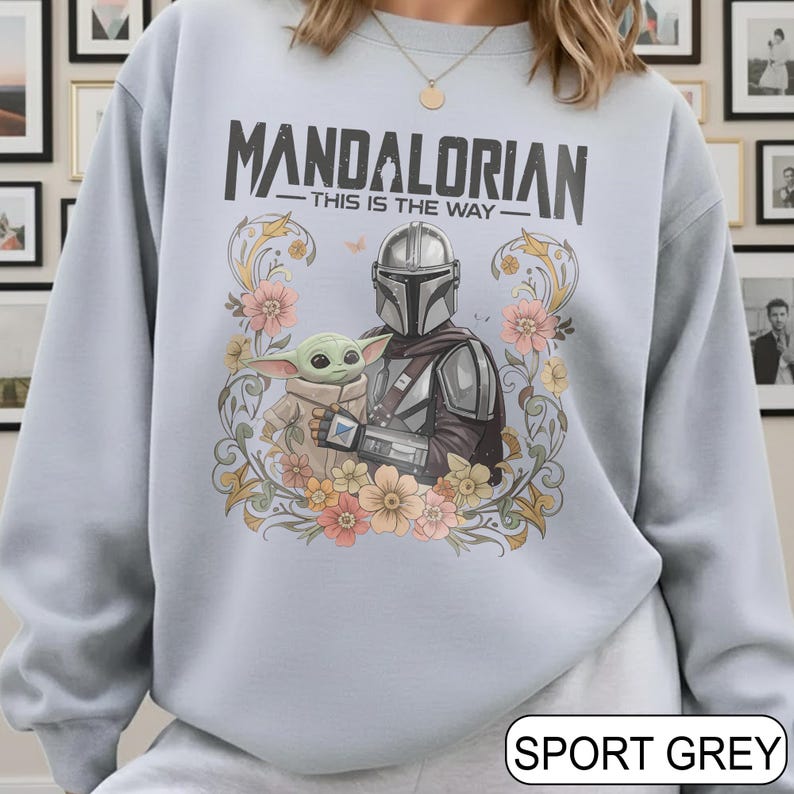 May include: Light blue sweatshirt featuring a graphic of The Mandalorian and Grogu, surrounded by floral elements. The text "MANDALORIAN - THIS IS THE WAY" is printed above the characters. The sweatshirt is labeled "SPORT GREY" at the bottom.