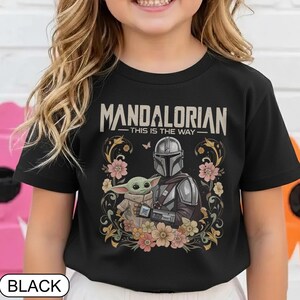 May include: Black t-shirt featuring a graphic of the Mandalorian and Grogu surrounded by flowers. The text "MANDALORIAN - THIS IS THE WAY" is printed across the top. The shirt is a crew neck.