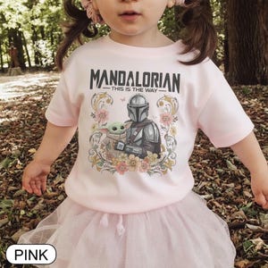May include: Pink t-shirt featuring the text "MANDALORIAN - THIS IS THE WAY" above an illustration of the Mandalorian and Grogu surrounded by floral elements. The shirt is worn by a child and paired with a pink tutu.
