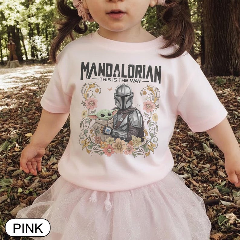 May include: Pink t-shirt featuring the text "MANDALORIAN - THIS IS THE WAY" above an illustration of the Mandalorian and Grogu surrounded by floral elements. The shirt is worn by a child and paired with a pink tutu.