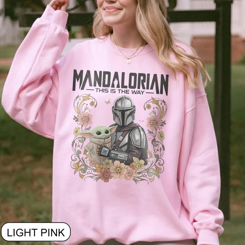 May include: Light pink sweatshirt featuring the text "MANDALORIAN - THIS IS THE WAY" in black. The graphic includes an illustration of the Mandalorian and Grogu surrounded by floral elements. The sweatshirt is a solid light pink color.