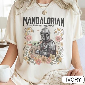 May include: Ivory-colored t-shirt featuring the text "MANDALORIAN" above "THIS IS THE WAY." The design includes an illustration of the Mandalorian and Grogu surrounded by flowers. The shirt is a short-sleeved, crew neck style.