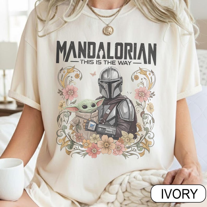 May include: Ivory-colored t-shirt featuring the text "MANDALORIAN" above "THIS IS THE WAY." The design includes an illustration of the Mandalorian and Grogu surrounded by flowers. The shirt is a short-sleeved, crew neck style.