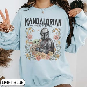 May include: Light blue sweatshirt featuring a graphic of The Mandalorian and Grogu surrounded by flowers. The text "MANDALORIAN - THIS IS THE WAY" is printed above the characters. The sweatshirt is a light blue color.