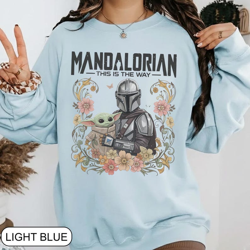 May include: Light blue sweatshirt featuring a graphic of The Mandalorian and Grogu surrounded by flowers. The text "MANDALORIAN - THIS IS THE WAY" is printed above the characters. The sweatshirt is a light blue color.