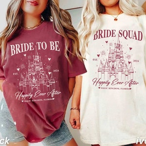 K&ouml;nnte beinhalten: Zwei T-Shirts mit einem wei&szlig;en Motiv von Schloss Aschenputtel auf einem burgunderfarbenen und elfenbeinfarbenen Hintergrund. Der Text auf dem burgunderfarbenen Shirt lautet "Bride to Be" und "Happily Ever After" mit dem Jahr "2024" und "EST.". Der Text auf dem elfenbeinfarbenen Shirt lautet "Bride Squad" und "Happily Ever After" mit dem Jahr "2024" und "EST."