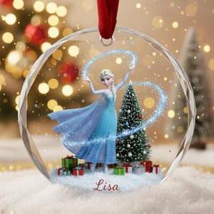 Puede incluir: Un adorno navideño redondo y transparente con un personaje de dibujos animados con un vestido azul y una capa fluida, de pie frente a un árbol de Navidad decorado. El adorno tiene una cinta roja para colgar y el nombre "Lisa" está impreso en la parte inferior.