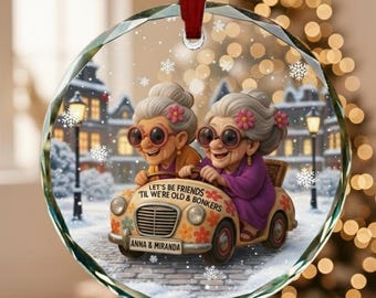 May include: A clear, round Christmas ornament with a red ribbon. The ornament features a cartoon illustration of two elderly women in a vintage car with the text "LET'S BE FRIENDS 'TIL WE'RE OLD & BONKERS" and "ANNA & MIRANDA". The background shows a snowy village.