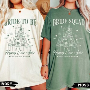 K&ouml;nnte beinhalten: Zwei T-Shirts mit einer gr&uuml;nen Umrandung von Cinderellas Schloss, Text, der "Bride to Be" und "Bride Squad" sagt, und die Worte "Happily Ever After" mit dem Text "Magic Kingdom, Florida" darunter. Das Shirt auf der linken Seite ist elfenbeinfarben und das Shirt auf der rechten Seite ist moosfarben.