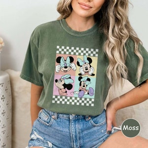 May include: A green t-shirt featuring a vintage-style graphic of Minnie Mouse in four different poses. The graphic is surrounded by a checkered border.