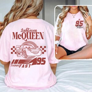 May include: Pink t-shirt featuring a red graphic of Lightning McQueen from the Cars film. The graphic includes the text "Lightning McQueen", "Think Fast", and "95".