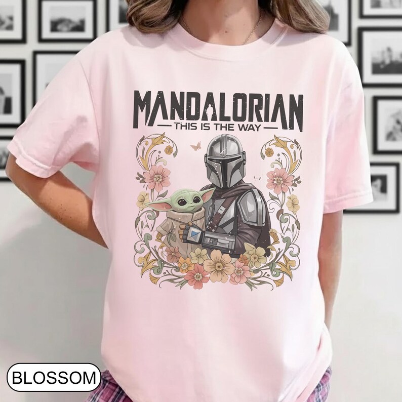 May include: Light pink t-shirt featuring a graphic of The Mandalorian and Grogu surrounded by floral elements. The text "MANDALORIAN - THIS IS THE WAY" is printed above the characters. The shirt has short sleeves.