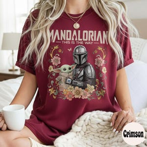 May include: Burgundy t-shirt featuring a graphic of The Mandalorian and Grogu surrounded by flowers. The text "MANDALORIAN - THIS IS THE WAY" is printed above the characters. The shirt has short sleeves and a crew neck.