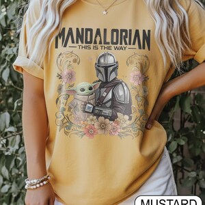 May include: Mustard-colored t-shirt featuring the text "MANDALORIAN" above an illustration of the Mandalorian and Grogu, surrounded by floral elements. The phrase "THIS IS THE WAY" is below the title. The shirt is a short-sleeved crew neck.