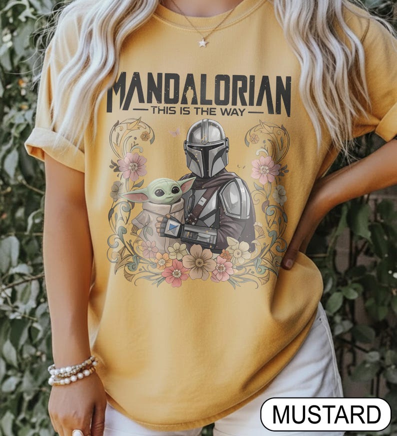 May include: Mustard-colored t-shirt featuring the text "MANDALORIAN" above an illustration of the Mandalorian and Grogu, surrounded by floral elements. The phrase "THIS IS THE WAY" is below the title. The shirt is a short-sleeved crew neck.