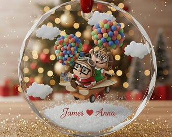 Puede incluir: Una bola de Navidad redonda y transparente con una cinta roja. En el interior, una pareja con equipo de aviador monta en un avión de madera, levantado por globos de colores. También aparecen nubes blancas y los nombres "James ❤️ Anna".