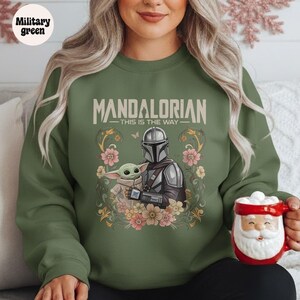 May include: Military green sweatshirt featuring a graphic of The Mandalorian and Grogu surrounded by flowers. The text "MANDALORIAN - THIS IS THE WAY" is printed above the characters. A red and white Santa mug is held in the person's hand.