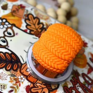 May include: A bright orange pumpkin-shaped accessory with a textured knit pattern, featuring a small brown stem. It is attached to a circular base that sits atop a white surface decorated with a cartoon ghost wearing glasses and autumn leaves.