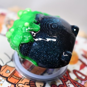 May include: A dark blue, glittery, rounded object resembling a cauldron with a glossy finish. A bright, neon green, textured substance that looks like bubbling liquid is dripping over one side of the dark blue surface, creating a high contrast.