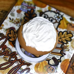 May include: A round, tan-colored container topped with a thick layer of white, textured cream. The container rests on a clear circular base, positioned over a surface decorated with a repeating pattern of autumn leaves, pumpkins, and books.
