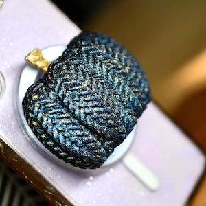 May include: A circular button featuring a dark blue and black braided herringbone pattern with subtle gold metallic flecks throughout the surface. The button has a raised, textured appearance and is mounted on a smooth, light-colored circular base.