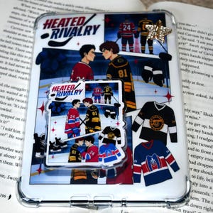 May include: A clear phone case featuring a hockey-themed design with the text "HEATED RIVALRY." The case includes illustrations of hockey players, jerseys, and a star-shaped embellishment. The design incorporates red, blue, black, and gold colors.