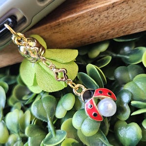 May include: A gold-colored phone charm featuring a ladybug design with red and black enamel and a pearl accent. The charm is attached to a gold chain and a decorative gold-colored flower. The background is a mix of green foliage and wood.