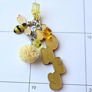 May include: A charm with a bee, pom-pom, and the word "HONEY" in gold. The bee has black and yellow stripes. The charm also includes yellow and clear beads. The background is a calendar.