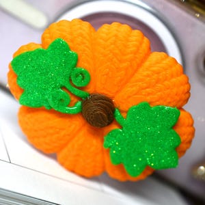 May include: A bright orange pumpkin figurine with a textured, ribbed surface. It features two vibrant green, glittery leaves attached to the top, along with a small, dark brown, coiled stem in the center. The object has a rounded, plump shape.