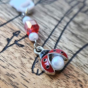 May include: A ladybug charm with red enamel wings and a pearl bead body. The charm is attached to a chain with a red faceted bead and white pearl beads. The ladybug is silver-toned.
