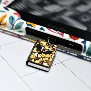 May include: A small, book-shaped charm with a black bat design and gold accents hangs near a Kindle e-reader. The charm has a silver-colored loop. The Kindle is in a floral patterned case with a white, pink, blue, and orange design.