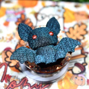 May include: A dark blue, textured bat figurine with two small, round, metallic red eyes. The bat is mounted on a circular, clear acrylic base that contains small, reflective copper-colored flakes embedded within the material.