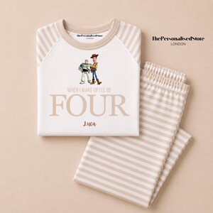 May include: A white long-sleeve pajama set with beige striped sleeves and pants. The shirt features Buzz Lightyear and Woody, the text "WHEN I WAKE UP I'LL BE FOUR", and the name "Luca". The brand "ThePersonalisedStore LONDON" is printed on the shirt.