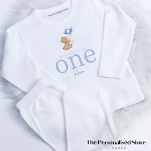 May include: White long-sleeved onesie with blue text that says "When I wake up I'll be one" and the name Rocco. A small illustration of a brown bear holding blue balloons is above the text.