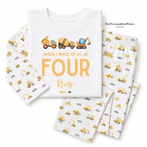 May include: A white long-sleeve pajama set featuring a repeating pattern of yellow construction vehicles. The shirt displays the text "WHEN I WAKE UP I'LL BE FOUR" in orange, the name "Reign" in script, and a small shovel icon.
