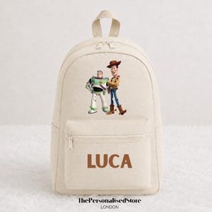 Op de afbeelding: Een beige canvas rugzak met een grafische print van Buzz Lightyear en Woody uit Toy Story op het hoofdvak. Het voorvak toont de naam LUCA in bruine hoofdletters. De tas is voorzien van een handvat aan de bovenkant en ritssluitingen.