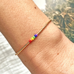 Könnte beinhalten: Ein zartes Armband mit einem durchgehenden Strang aus kleinen, metallisch goldfarbenen zylindrischen Perlen. Eine einzelne, mittig platzierte Abfolge von sechs winzigen, runden, bunten Perlen in Rot, Orange, Gelb, Grün, Blau und Violett.