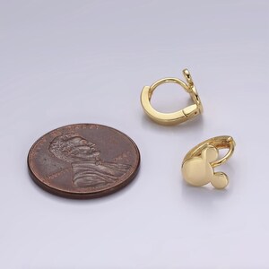 Op de afbeelding: Twee goudkleurige oorringen met een Mickey Mouse-ontwerp, naast een penny. De oorbellen hebben een gladde, gepolijste afwerking en een scharniersluiting. De Mickey Mouse-kop heeft een ronde vorm met twee kleine, ronde oren.