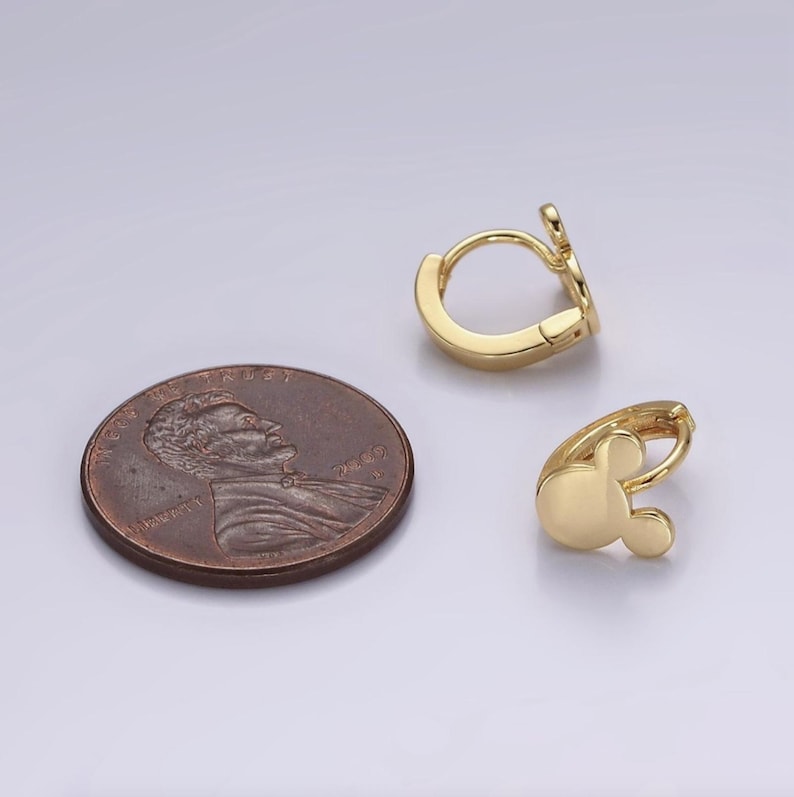 Op de afbeelding: Twee goudkleurige oorringen met een Mickey Mouse-ontwerp, naast een penny. De oorbellen hebben een gladde, gepolijste afwerking en een scharniersluiting. De Mickey Mouse-kop heeft een ronde vorm met twee kleine, ronde oren.