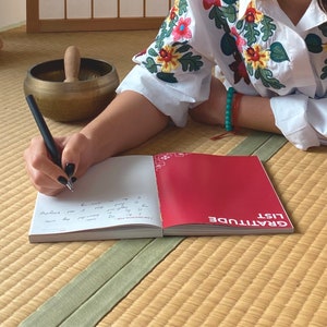 Może przedstawiać: Osoba pisze w czerwono-białym dzienniku wdzięczności czarnym długopisem. Na okładce dziennika widnieje napis "Gratitude List".
