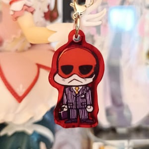 May include: A red acrylic charm featuring a cartoon character in a suit with a red head and sunglasses. The charm is attached to a silver-colored clasp with a small wing design.