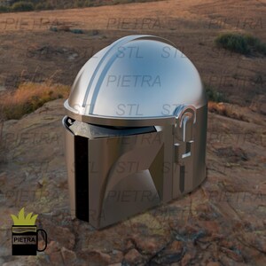 Puede incluir: Un casco plateado con un diseño en relieve en la parte delantera y una visera negra. El casco tiene una parte superior plateada con una línea en relieve en el centro. El casco está sentado sobre una superficie rocosa.