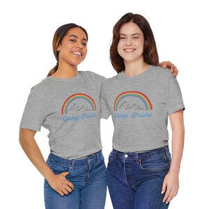 May include: Two people wearing grey t-shirts with a rainbow mountain and the text "Camp Pride" printed on the front. They are both smiling and looking at the camera.